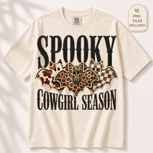 May include: A cream-colored t-shirt featuring the black text SPOOKY COWGIRL SEASON. Between the words are three bat illustrations with cow print, leopard print, and checkered patterns, accented with small turquoise-colored circular studs.