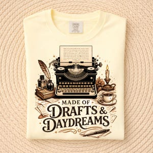 May include: Cream-colored t-shirt featuring a vintage typewriter illustration with a feather quill, inkwell, and coffee cup. The text "MADE OF DRAFTS & DAYDREAMS" is displayed below the typewriter. The shirt is folded.