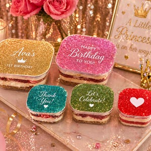May include: A selection of square desserts with glitter lids in gold, pink, teal, green, and red. The lids have text: "Ava's 1st Birthday", "Happy Birthday to You!", "Thank You!", and "Let's Celebrate!" A heart is on the red lid. The desserts are on a tray.