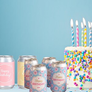 May include: A birthday celebration scene featuring several aluminum cans with "Happy Birthday!" labels, alongside a decorated cake with lit candles. The cans have floral and solid color designs.