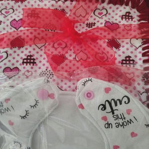 May include: A gift set featuring a white and pink heart-patterned blanket with a red crocheted border and a sheer red ribbon. Includes two white bibs with eyelashes and the text "I woke up this cute."