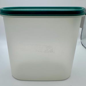 May include: A rectangular, off-white plastic food storage container with a dark green lid. The container has a slightly frosted appearance and a smooth, clean design. The lid is rectangular and fits snugly on top.