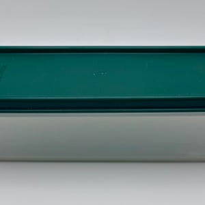 Può includere: Un contenitore rettangolare per alimenti con un coperchio verde scuro. Il contenitore è traslucido, consentendo la visibilità del contenuto. Il coperchio ha angoli arrotondati e si adatta perfettamente alla base. Il contenitore è progettato per la conservazione degli alimenti.