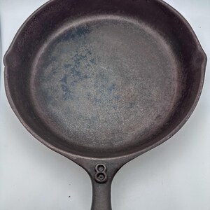 以下が含まれることがあります： よく使い込まれた鋳鉄製のフライパンで、濃く古びた外観をしています。 フライパンは丸みを帯びた形状で、長い柄と片側に小さな注ぎ口が付いています。 ハンドルにはサイズを示す数字の「8」がエンボス加工されています。