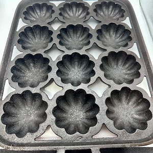 May include: A cast iron madeleine pan with a rectangular shape and two handles. The pan features twelve shell-shaped molds, perfect for baking the classic French madeleine cakes. The pan is dark gray and has a rustic, well-used appearance.