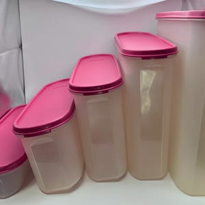 Pode incluir: Um conjunto de cinco recipientes de plástico transparente para armazenamento de alimentos com tampas rosa. Os recipientes variam em tamanho, com um recipiente alto e retangular à direita e recipientes menores e angulados à esquerda. As tampas são de um tom rosa mais escuro.