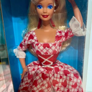 Puede incluir: Una muñeca rubia con ojos azules y lápiz labial rojo, vestida con un vestido rojo y blanco con estampado de corazones. La muñeca tiene una diadema roja y está en una caja azul. El vestido tiene un escote cuadrado y un corpiño estilo corsé.