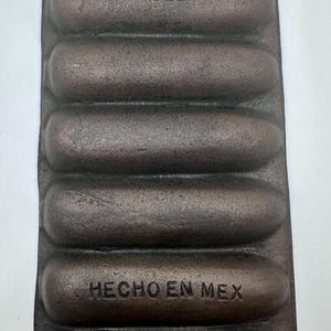 May include: A cast iron cornbread pan with six individual molds. The pan is dark brown with the word "PAGOEL" and "HECHO EN MEX" embossed on the surface. It has a handle for easy handling.