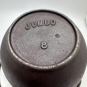 May include: A cast iron pot with a dark, textured surface. The word "JUMBO" is embossed on the lid, along with the number "8". The pot has a rounded shape and a metal handle. This cookware is suitable for cooking.