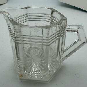 Puede incluir: Una jarra de crema de vidrio transparente, de diseño octogonal. La jarra tiene un asa y un pico vertedor. El vidrio está grabado con un patrón de estrella. Un objeto decorativo para la mesa.