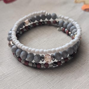 May include: A multi-strand wrap bracelet featuring a combination of white seed beads, faceted grey beads, small dark red beads, and metallic silver spacers. The design includes a single translucent peach-colored crystal bead as a central accent.