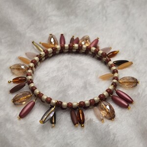 May include: A multi-strand wrap bracelet featuring alternating cream and copper-colored glitter beads. The bracelet is adorned with dangling charms in various shapes, including faceted amber crystals, matte mauve beads, and smooth brown beads.