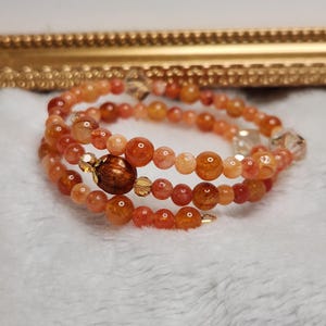May include: A multi-strand wrap bracelet featuring round, translucent orange and reddish-brown gemstone beads. The design includes a larger, dark brown oval-shaped focal bead and smaller, clear faceted crystal accents interspersed throughout the piece.