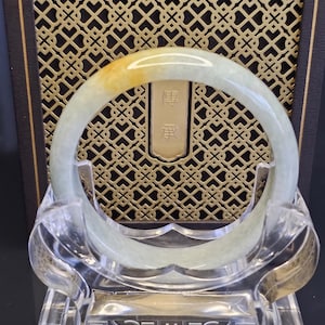 May include: A smooth, circular jade bangle featuring a pale green hue with a distinct golden-yellow patch. The bangle is displayed on a clear acrylic stand in front of a dark brown box with a gold-colored lattice pattern and Chinese characters.