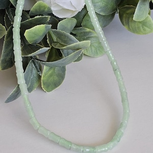 Pode incluir: Um colar de contas verde claro com um design delicado. O colar é feito de pequenas contas retangulares enfiadas juntas. As contas têm uma aparência ligeiramente translúcida. O colar é exibido em um fundo neutro com folhagem verde e flores brancas.