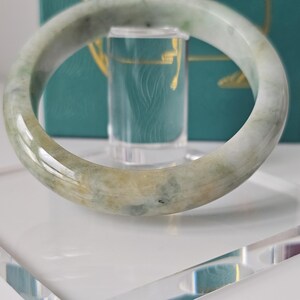 May include: A smooth, polished jade bangle bracelet featuring a translucent appearance with mottled light green, white, and soft yellow-brown color variations throughout the circular stone band, displayed on a clear acrylic stand.