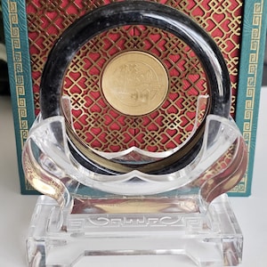 May include: A dark jade bangle bracelet displayed on a clear acrylic stand. The bracelet is set against a red and gold patterned background within a teal box. A gold coin is visible inside the bracelet.