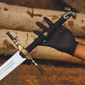 May include: A detailed image of a sword with a silver blade and gold accents. The hilt is wrapped in black material and features a decorative silver lion's head. The sword is held by a gloved hand against a backdrop of wooden logs.