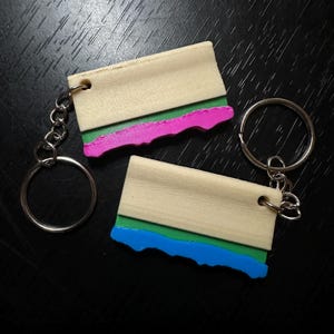 May include: Two wooden keychains with a light wood finish, each featuring a green and a pink or blue accent. The keychains are attached to silver key rings. The background is a dark surface.