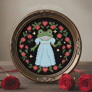 Könnte beinhalten: Ein gerahmtes Kreuzstichbild mit einem Frosch in einem hellblauen Kleid, umgeben von roten Rosen und Herzen auf schwarzem Hintergrund. Der Rahmen ist goldfarben, und der Text "VelourPeachXStitch" ist oben sichtbar. Rote Rosen sind auf einer Holzoberfläche angeordnet.