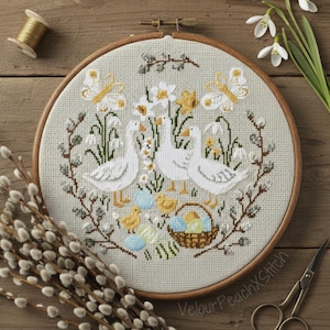 Könnte beinhalten: Ein Kreuzstich-Stickrahmen mit einer Frühlingsszene, die drei weiße Gänse, Schmetterlinge und einen Korb mit Ostereiern zeigt. Das Design umfasst gelbe Narzissen und Schneeglöckchen auf hellgrünem Hintergrund. Der Rahmen liegt auf einer Holzoberfläche.