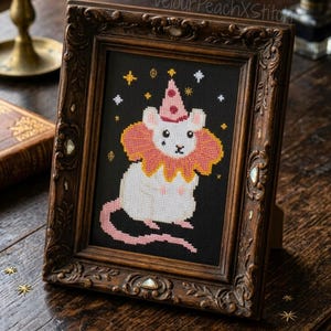 Peut inclure: Une œuvre d'art au point de croix encadrée représentant une souris fantaisiste avec un chapeau de fête et un col à volants, sur fond noir avec des étoiles. Le cadre est en bois foncé orné de détails décoratifs. L'œuvre est exposée sur une surface en bois.