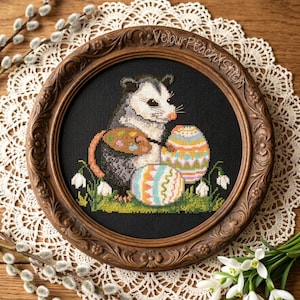 Könnte beinhalten: Ein gerahmtes Kreuzstichbild mit einem Opossum mit Ostereiern und einem Korb. Das Kunstwerk ist vor einem schwarzen Hintergrund in einem detaillierten braunen Holzrahmen gerahmt. Der Text "Velour Peach x Stitch" ist auf dem Rahmen zu sehen.