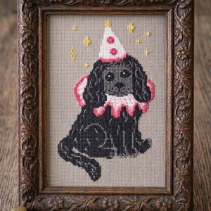 Könnte beinhalten: Kreuzstich-Kunst eines schwarzen Hundes mit Partyhut und Rüschenkragen, gerahmt in einem detaillierten Holzrahmen. Gestickte Sterne umgeben den Hund. Das Kunstwerk ist mit "VelourPeachXStitch" signiert.