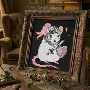 May include: Cross-stitch art of a white mouse in knight's armor, holding a sword and shield, set against a black background. The framed piece has a decorative, ornate brown frame and is displayed on a table. The text "VelourPeachXStitch" is visible.