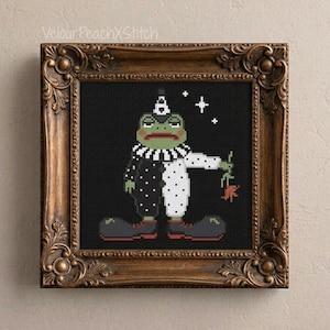 May include: A cross-stitch embroidery featuring a green frog dressed as a clown in a black and white polka-dot outfit with a ruffled collar, holding a small red flower, framed in an ornate antique bronze-colored frame with text VelourPeachXStitch.