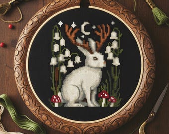 May include: Cross-stitch embroidery of a white rabbit with deer antlers, surrounded by white flowers and red mushrooms, set against a black background. The design is framed in a wooden hoop with carved details. The text "Velour Peach XStitch" is at the bottom.