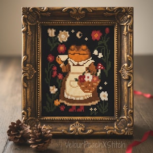 Puede incluir: Arte de punto de cruz en un marco dorado decorativo. La obra presenta un personaje de rana con un delantal y sosteniendo una taza de té y una cesta de flores. El fondo es negro con detalles florales. El marco tiene detalles ornamentados.