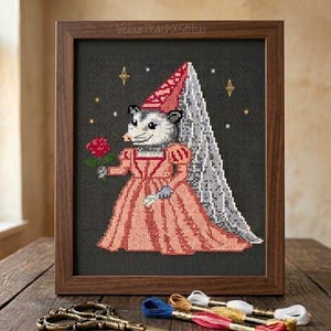 Könnte beinhalten: Kreuzstich-Kunstwerk in einem Holzrahmen, das ein Opossum in einem rosa Kleid und einem roten Hut zeigt, das eine rote Rose hält. Das Opossum hat ein grau-weißes Gesicht und einen grauen Schleier. Der Hintergrund ist dunkelgrau mit goldenen Sternen.