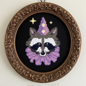 Könnte beinhalten: Ein Kreuzstich-Kunstwerk eines Waschbären mit lila Partyhut und Kragen, vor schwarzem Hintergrund. Das Kunstwerk ist in einem detaillierten, verzierten, ovalen, bronzefarbenen Rahmen gerahmt. Der Text "VelourPeachXStitch" befindet sich unten.