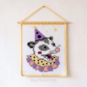 Könnte beinhalten: Ein rechteckiger Wandbehang aus Häkelarbeit mit einem Opossum im Kreuzstich-Stil, das einen violetten Partyhut mit Punkten und einen gerüschten gelb-violetten Kragen trägt. Das Stück hängt an einem Holzstab mit einer gelben Garnaufhängung.