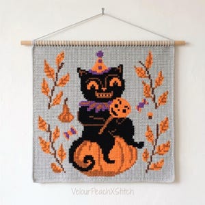 Könnte beinhalten: Ein quadratischer Häkel-Wandbehang an einem Holzstab mit einer schwarzen Katze, die einen orange-violetten Partyhut und Kragen trägt, auf einem großen orangefarbenen Kürbis sitzt, einen kleinen Kürbis hält und von orangefarbenen Herbstblättern umgeben ist.