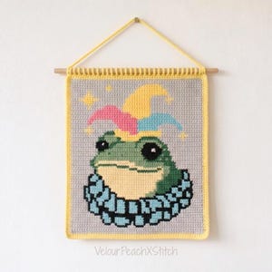 Könnte beinhalten: Ein rechteckiger Wandbehang aus Häkelarbeit mit einem grünen Frosch, der eine bunte Narrenkappe und einen blauen Rüschenkragen vor grauem Hintergrund trägt. Das Stück hängt an einem Holzstab mit gelber Kordel und enthält den Text VelourPeachXStitch.