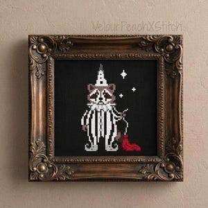 May include: A cross-stitch embroidery on black fabric featuring a raccoon dressed as a clown in a white striped suit and pointed hat. The raccoon holds a red string attached to a red puddle. The piece is encased in an ornate dark brown frame.