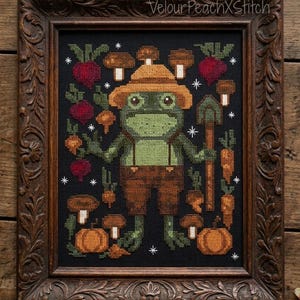 Könnte beinhalten: Kreuzstich-Kunstwerk eines Frosch-Gärtners mit Hut und Latzhose, der eine Schaufel und Karotten hält, umgeben von Pilzen, Kürbissen und Rüben. Das Kunstwerk ist in einem dunkelbraunen, verzierten Holzrahmen gerahmt. Der Text "VelourPeachXStitch" ist sichtbar.