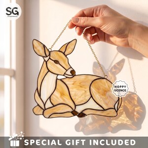May include: A stained glass suncatcher shaped like a resting deer, crafted from translucent beige and cream glass pieces with dark solder lines. The deer is suspended by a thin gold-toned metal chain, casting a soft shadow on the wall behind it.