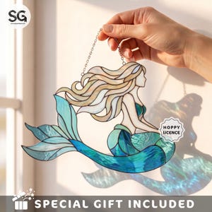 May include: A stained glass suncatcher shaped like a mermaid with flowing cream-colored hair and a tail featuring various shades of blue and teal glass. The piece hangs from a metal chain and includes a small white sticker labeled HOPPY LICENCE.