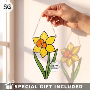 May include: A stained glass suncatcher shaped like a yellow daffodil with an orange center and green leaves. The piece features a metal chain for hanging and a small white label with the text HOPPY LICENCE attached to the lower right leaf.