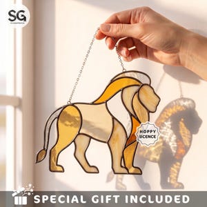 May include: A stained glass suncatcher shaped like a lion, featuring amber, yellow, and cream-colored glass segments held together by dark metal solder lines. The lion is suspended by a thin metal chain and includes a small tag reading HOPPY LICENCE.