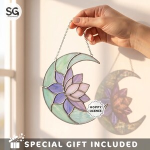 May include: A crescent moon-shaped stained glass suncatcher featuring a central lotus flower design with light purple and white petals, surrounded by pale green glass segments. The piece hangs from a silver-toned metal chain.