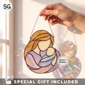 May include: A stained glass suncatcher depicting a stylized figure holding a baby, crafted with amber, lavender, and cream-colored glass pieces joined by dark metal solder lines. The piece hangs from a silver-toned metal chain.