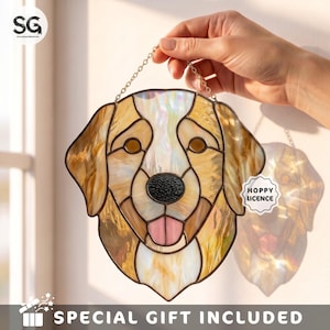 May include: A stained glass suncatcher shaped like a golden retriever head, featuring amber, white, and iridescent glass pieces with dark solder lines. The dog has a black textured nose and a pink tongue. It hangs from a thin metal chain.