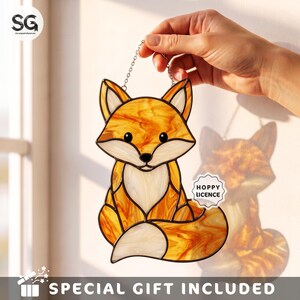 May include: A stained glass suncatcher shaped like a sitting fox, featuring orange and white marbled glass pieces held together by dark metal solder lines. The fox has black glass eyes and a nose, and a small white label reading HOPPY LICENCE.