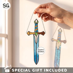 May include: A stained glass sword suncatcher featuring a blue textured blade, a gold and blue striped handle, and a green diamond accent at the crossguard. The piece is suspended by a silver metal chain and includes a white label reading HOPPY LICENCE.