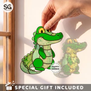 May include: A stained glass suncatcher shaped like a green crocodile with yellow eyes and a light green belly. The piece features dark metal solder lines outlining the segments. A small white sticker with the text HOPPY LICENCE is attached.
