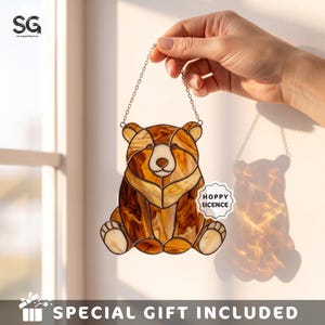 May include: A stained glass suncatcher shaped like a sitting bear, crafted from variegated brown and cream glass pieces joined by dark metal solder lines. It features a silver chain for hanging and a small white label reading HOPPY LICENCE.